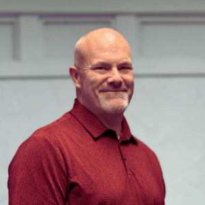 Dr. Michael Hartle in a red polo shirt standing in front of white wall. He is smiling.