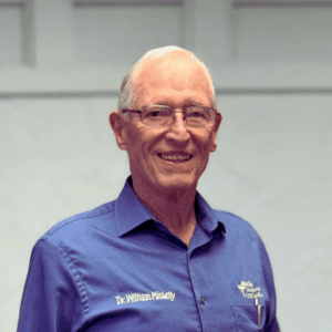 Dr. William (Bill) Miskelly in a blue button shirt standing in front of white wall. He is smiling. 