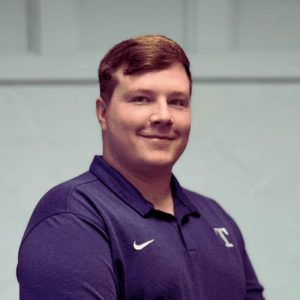 Dr. Nolan Stratton in a blue polo shirt standing in front of white wall. He is smiling.