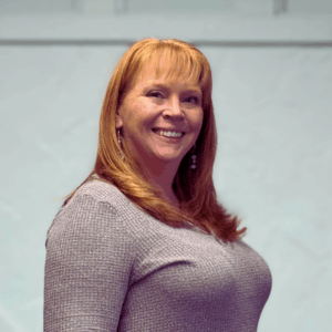 Office Manager, Holli Sauer in a gray sweater standing in front of white wall. She is smiling.