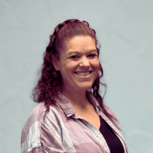 Massage Therapist, Shannon Boits in a pink and white checkered button shirt standing in front of white wall. She is smiling.